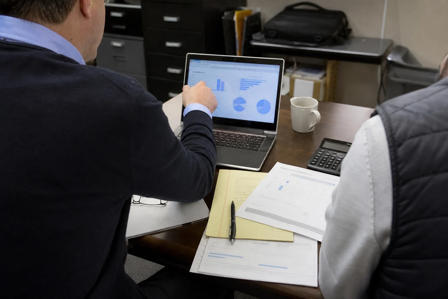 Advisor in a navy sweater points to charts on a laptop while a client in a gray vest looks on, with a legal pad, printed reports and a calculator on an office desk