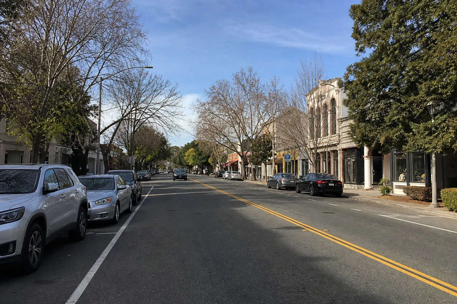 Wide view of a tree-lined downtown street with parked cars along both curbs, double yellow center lines and storefronts under a blue sky