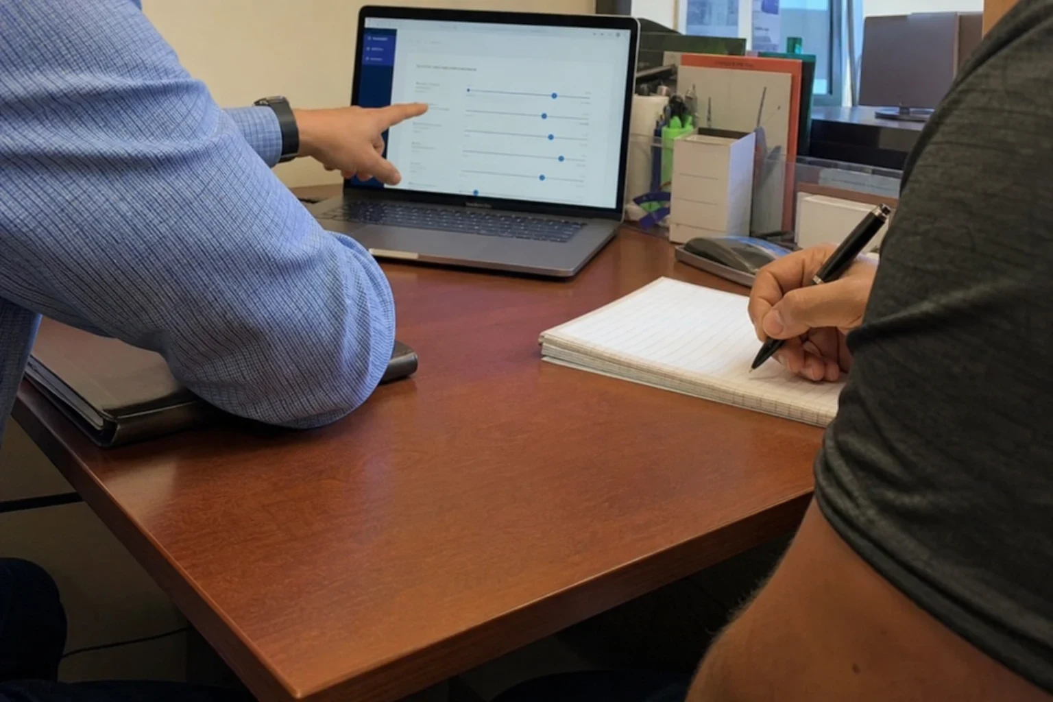 Agent in a checked shirt points at sliders on a laptop screen while a client in a gray shirt takes notes on a pad at a wooden office desk